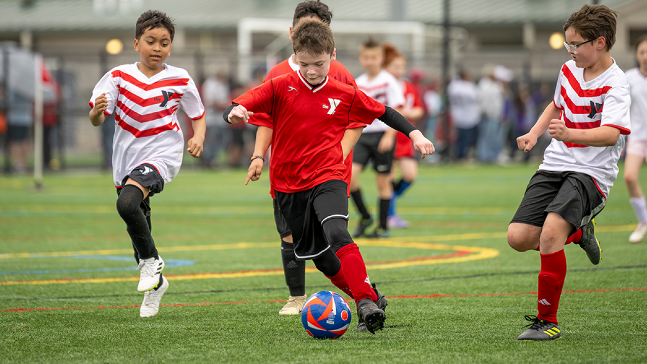 Soccer Home Hero August 2025 YMCA soccer game featuring a young boy in red dribbling the ball between defenders